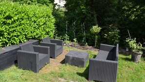 Mobiliario de mimbre exterior en un jardín con abundante follaje y un edificio al fondo.