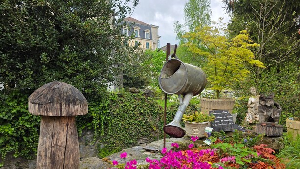 Ein Garten mit einer Skulptur aus einem Gießkannenmotiv, einer pilzförmigen Struktur und leuchtend rosa Blumen.