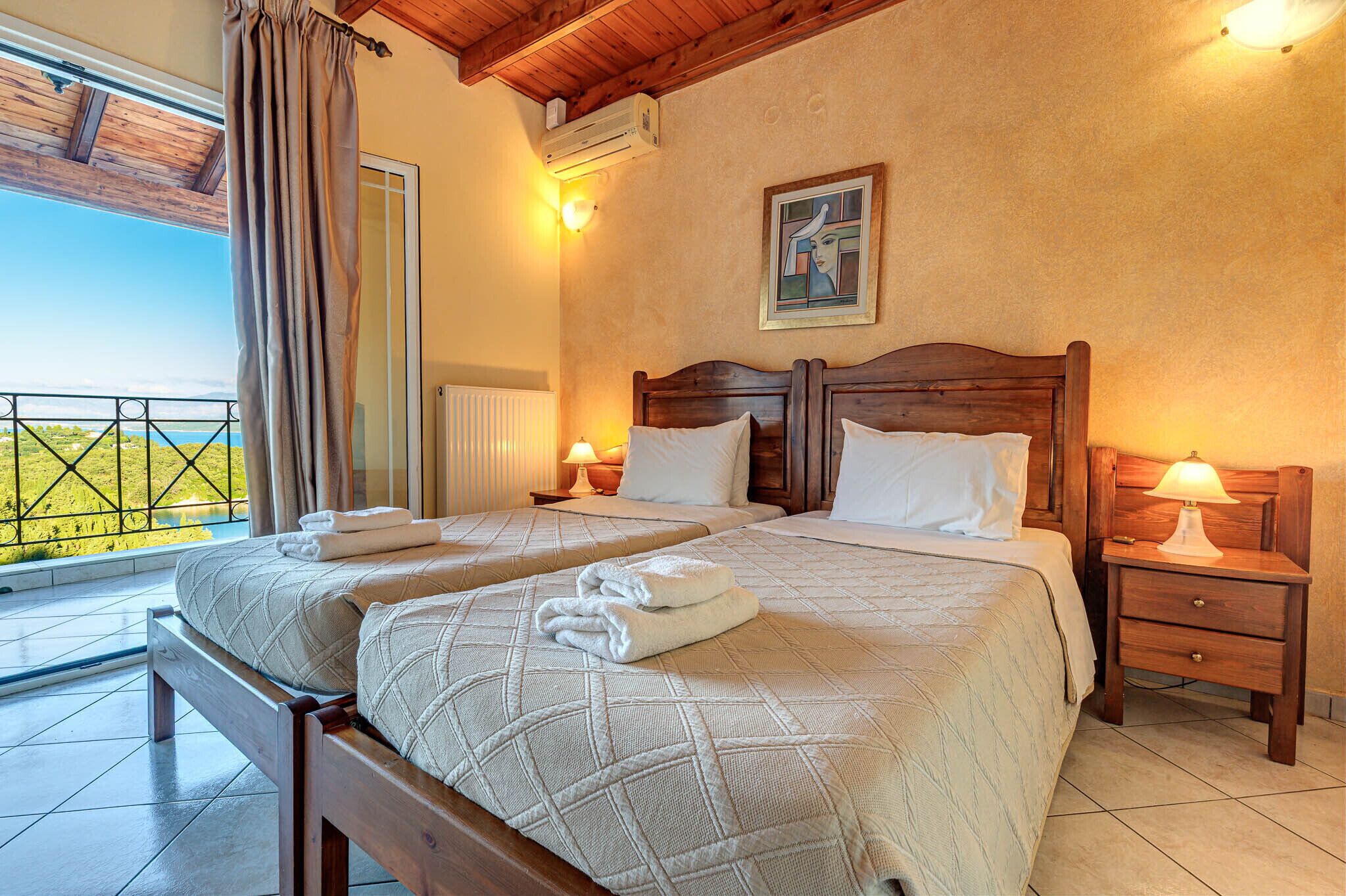 Une chambre avec deux lits, une tête de lit en bois, une table de chevet avec une lampe, et une vue sur la verdure par la fenêtre.