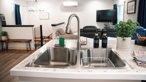 A modern kitchen with a stainless steel sink, a dining area with a bench, and a living room with a sofa and a TV. - Cozy Carriage House near Clemson - full kitchen, deck, fire pit, grill, and more (Central)