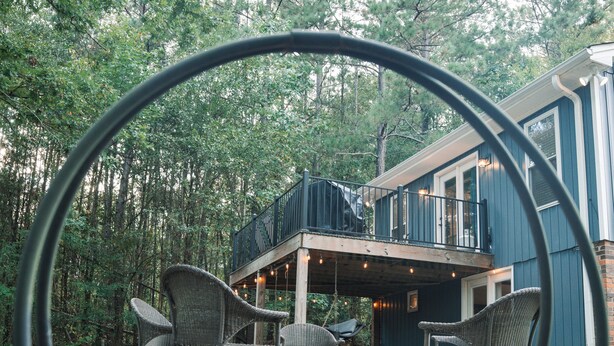 A wooden deck with wicker chairs, a stone fire pit, and a view of a house with a balcony. - Cozy Carriage House near Clemson - full kitchen, deck, fire pit, grill, and more (Central)