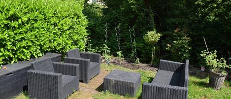 Outdoor wicker furniture in a garden setting with dense foliage and a building in the background.