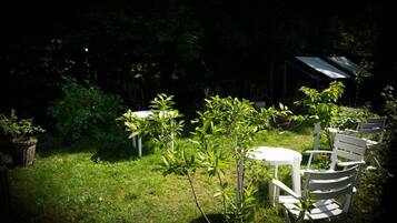Ein Garten mit einem weißen Tisch und Stühlen, umgeben von viel Grün und einem überdachten Bau.