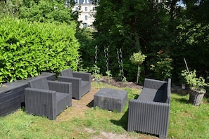 Outdoor wicker furniture in a garden setting with dense foliage and a building in the background. - La diligence de la poste – Furnished T2 at Domaine Du Bouleau with Air Conditioning (La Vôge-les-Bains)