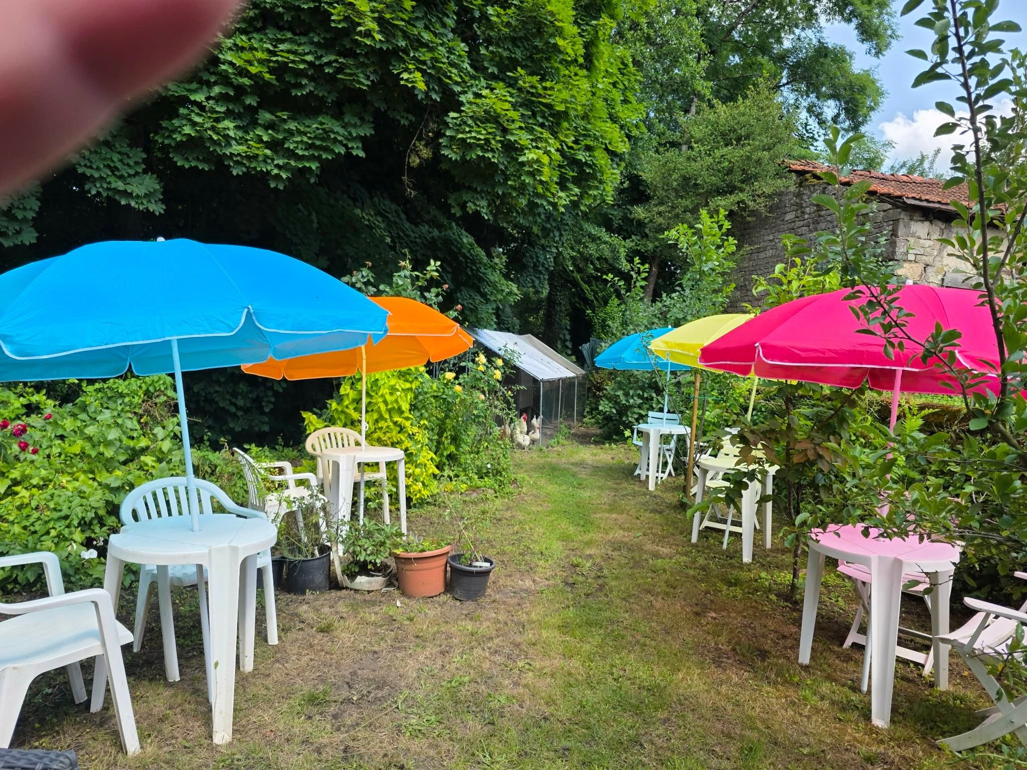 An outdoor seating area with multiple tables and chairs, surrounded by greenery and a stone wall.