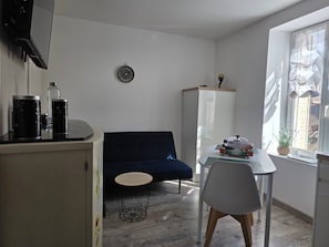 Et lille, moderne rum med en mørkeblå sofa, et rundt træbord, et hvidt skab og et vindue med udsigt til bygninger.
