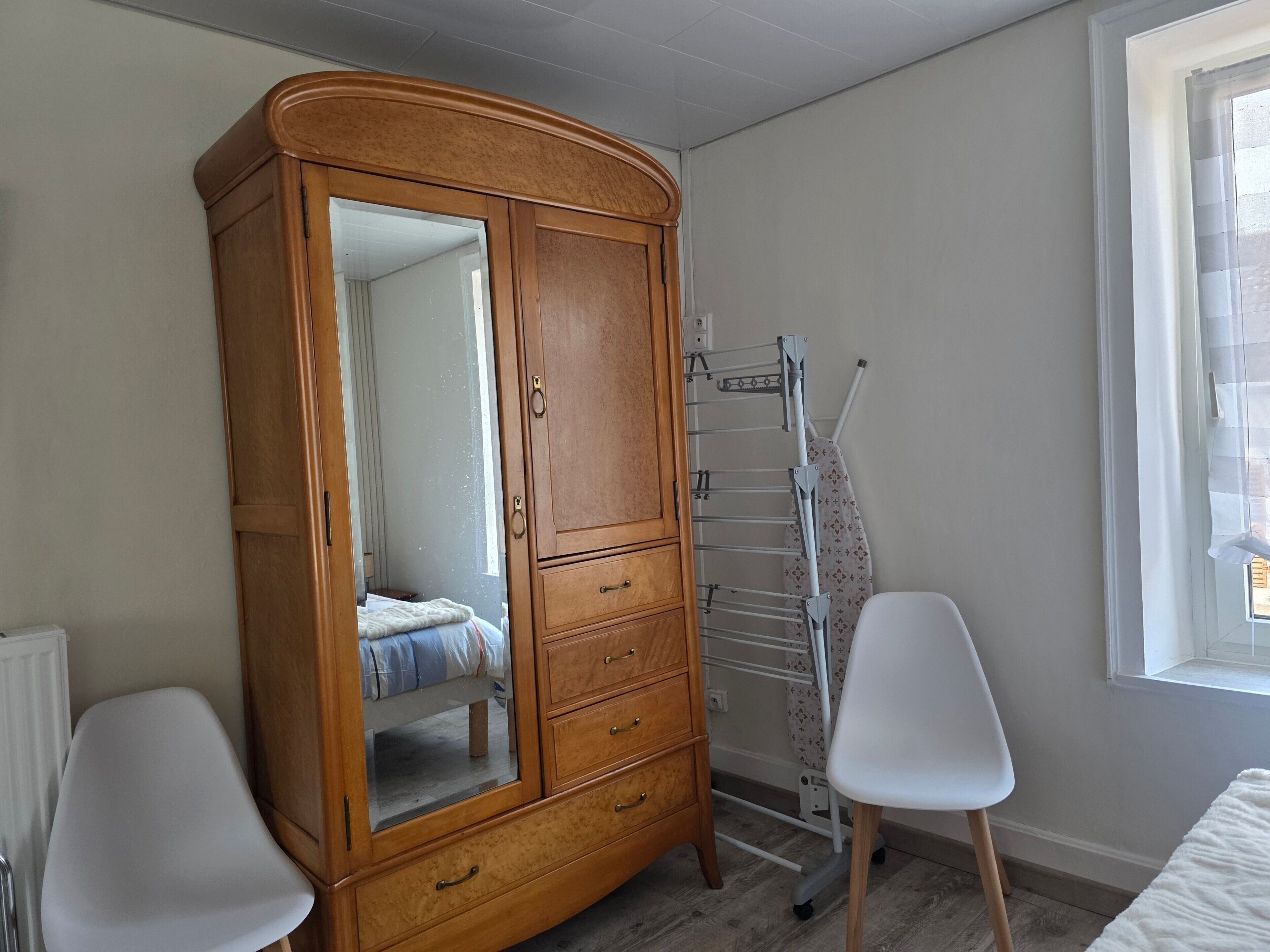 A bedroom with a wooden wardrobe, a mirror, a metal drying rack, a white chair, and a window.