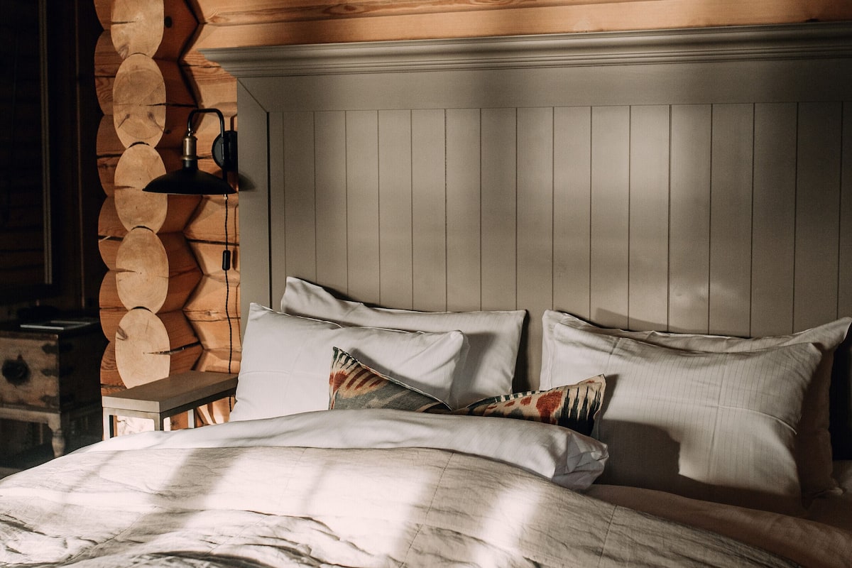 Interior de una cabaña de madera con una cama, una lámpara en la pared y una pared de troncos de madera.