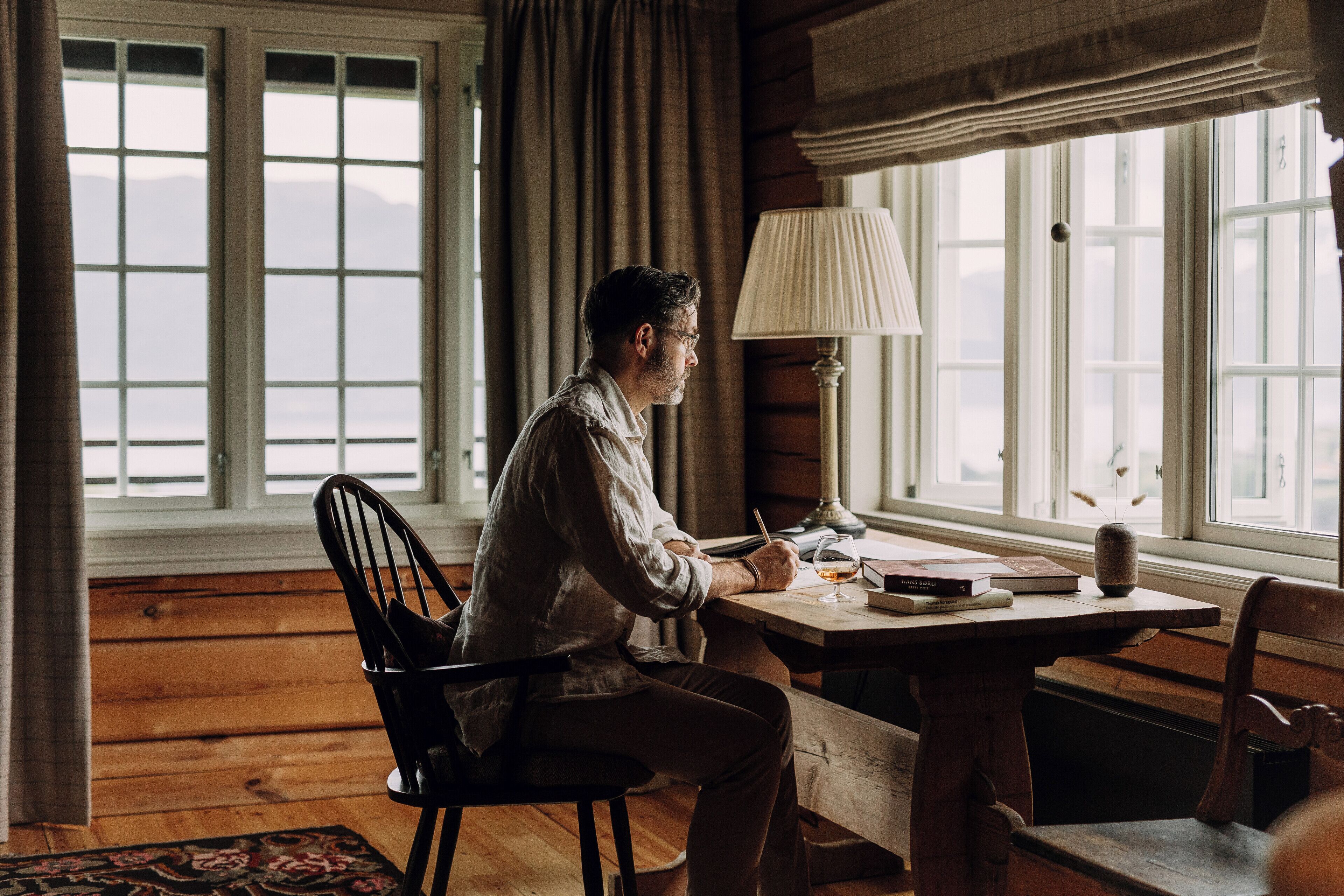 A man sitting at a wooden table in a room with large windows, writing in a notebook.