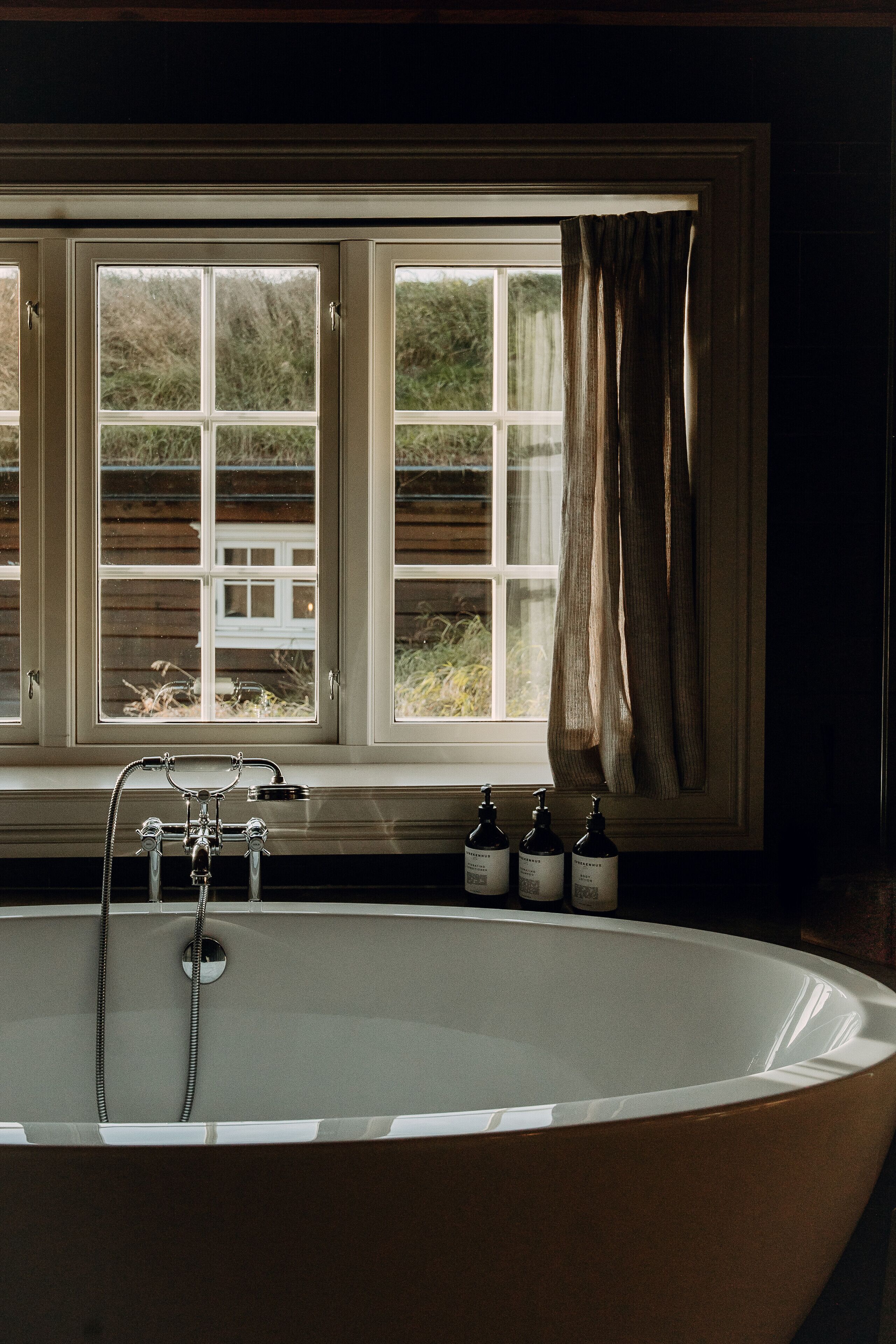 A bathroom with a freestanding bathtub, a window with curtains, and a shelf with toiletries.