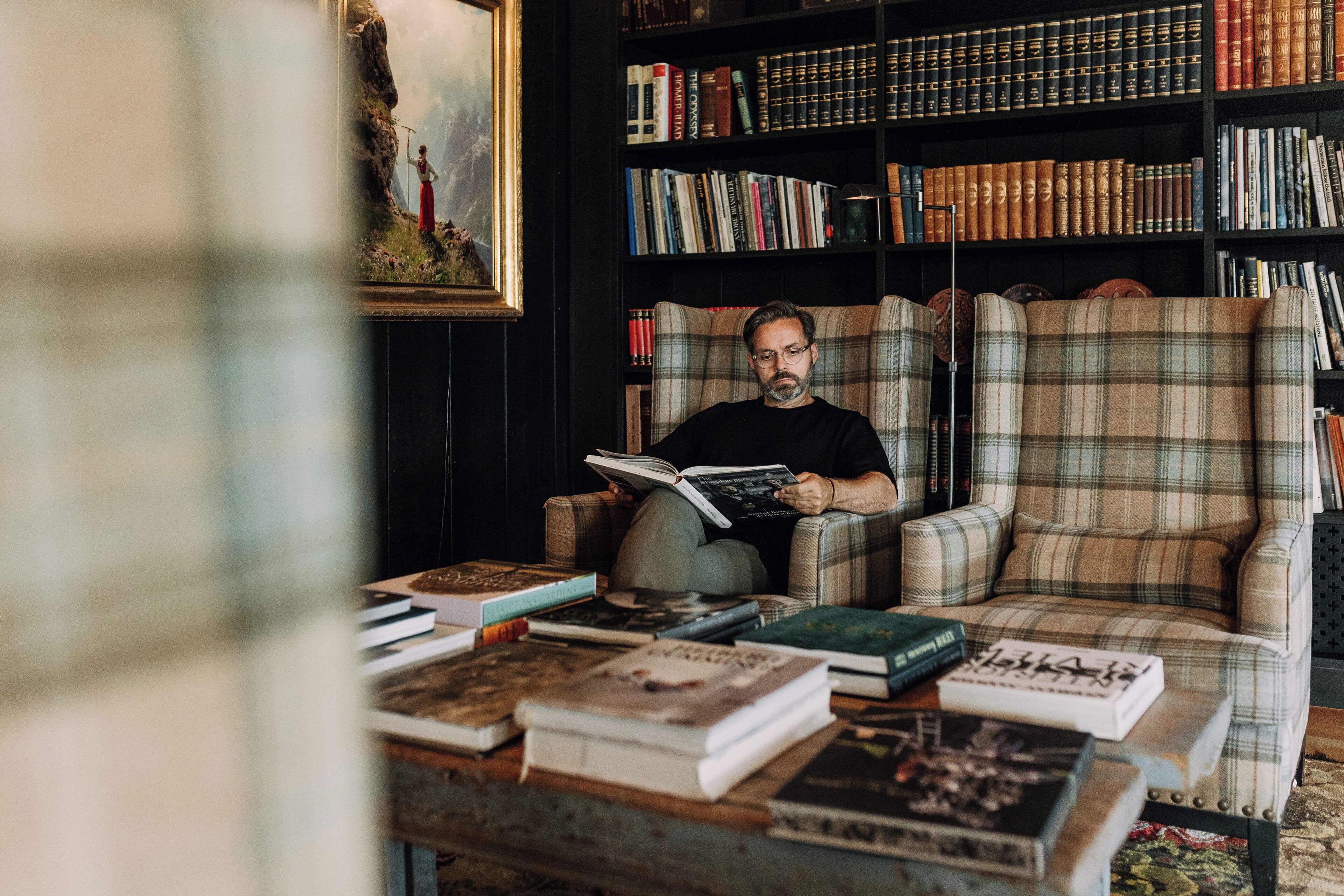 Une personne est assise dans une pièce, lisant un livre, avec une bibliothèque et un tableau accroché au mur.