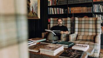 Une personne est assise dans une pièce, lisant un livre, avec une bibliothèque et un tableau accroché au mur.