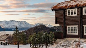 A log cabin with a stone chimney, wooden windows, and a view of snow-covered mountains. - Storfjord Hotel (Ålesund)