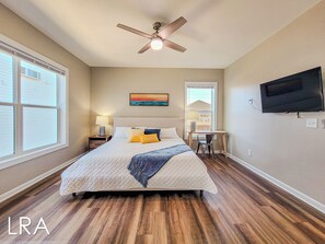 A bedroom with a bed, a desk, a chair, a ceiling fan, a TV, and a window with blinds. - Blue Crab Bungalow – Stay, Play, Repeat! (Surf City)