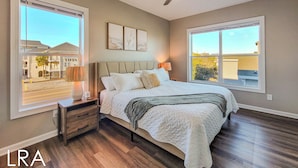 A bedroom with a bed, nightstands, a ceiling fan, and large windows. - Blue Crab Bungalow – Stay, Play, Repeat! (Surf City)