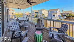 A covered outdoor deck with a table and chairs, a ceiling fan, and a view of residential buildings. - Blue Crab Bungalow – Stay, Play, Repeat! (Surf City)