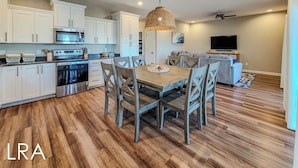 A modern kitchen with white cabinets, stainless steel appliances, and a dining area with wooden chairs and a table. - Blue Crab Bungalow – Stay, Play, Repeat! (Surf City)