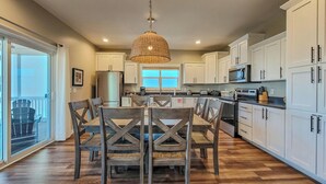 A modern kitchen with white cabinets, a dining table with chairs, and a hanging light fixture. - Blue Crab Bungalow – Stay, Play, Repeat! (Surf City)