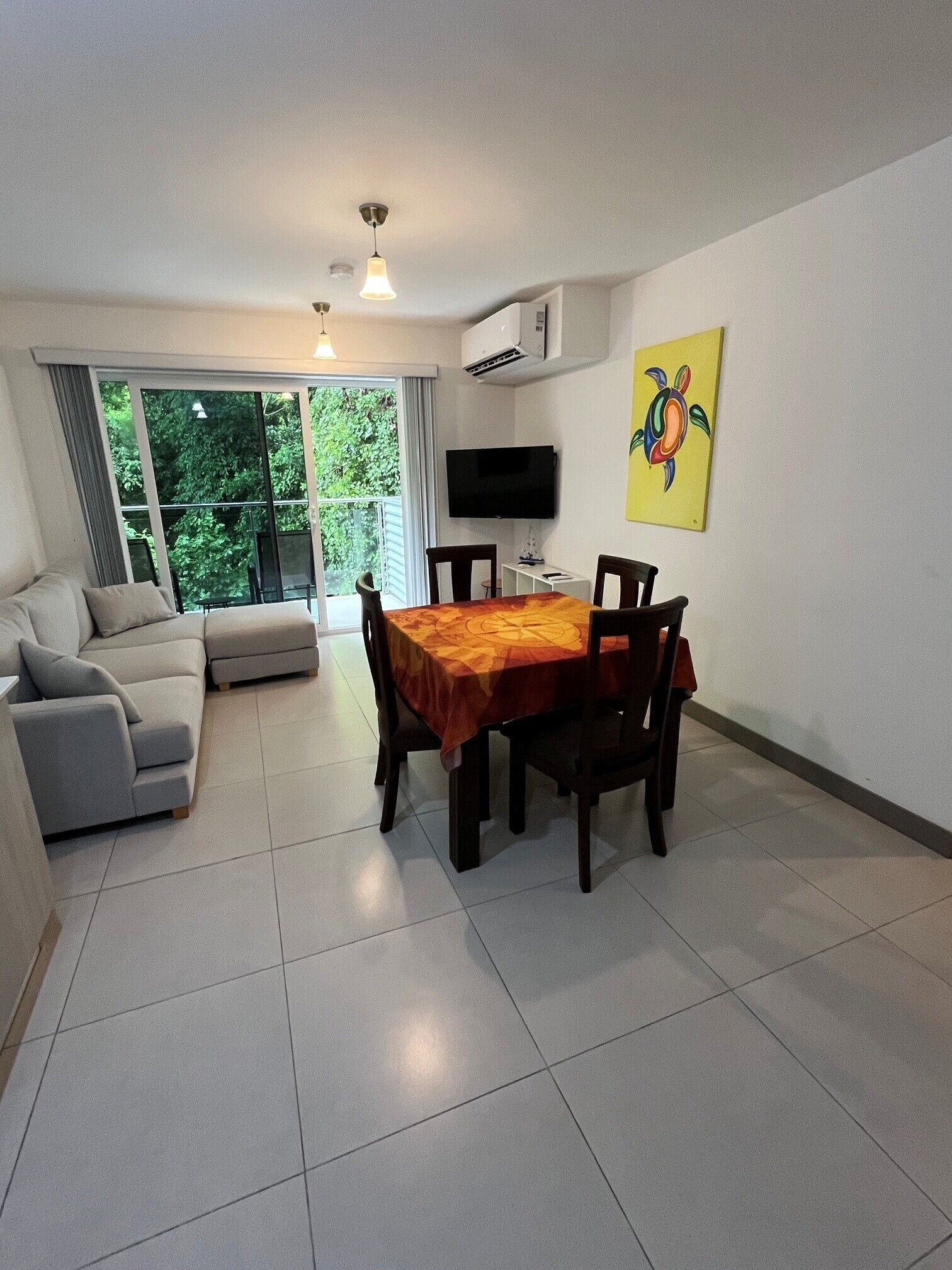 A living room with a sofa, a dining table with chairs, a flat-screen TV mounted on the wall, and a window with a view of greenery.