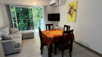 A living room with a sofa, a dining table with chairs, a flat-screen TV mounted on the wall, and a window with a view of greenery.