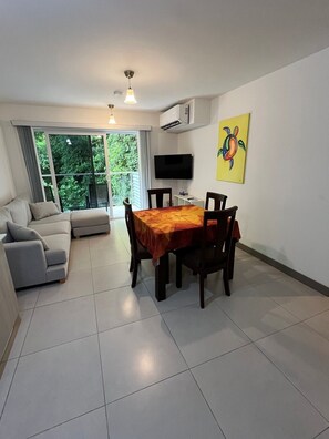 A living room with a sofa, a dining table with chairs, a flat-screen TV mounted on the wall, and a window with a view of greenery. - Stunning 2 Bedroom Apartment in Punta Esmeralda , Costa Rica (Tárcoles)
