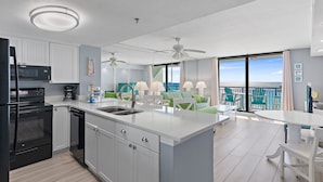 Una cocina moderna con armarios blancos, un horno negro y una isla grande con lavamanos. Hay un comedor con mesa y sillas blancas, y un balcón con vistas al océano.
