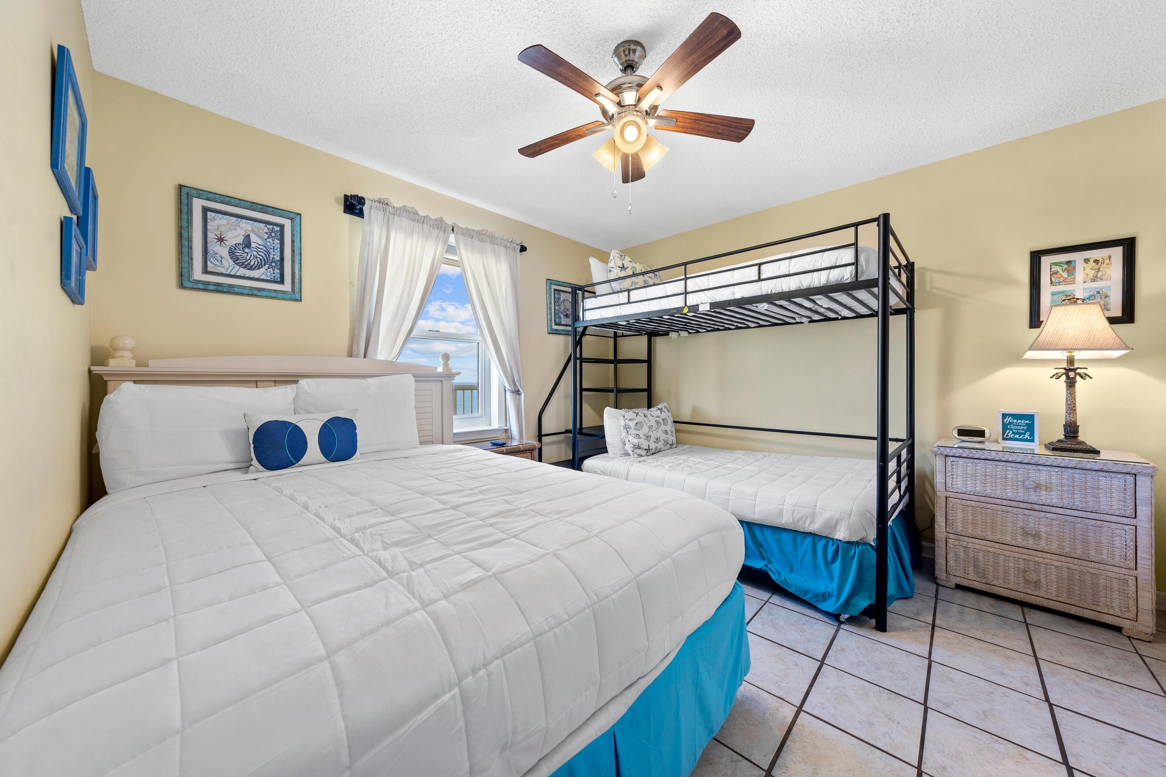 A bedroom with two bunk beds, a ceiling fan, a bedside table with a lamp, and a window with curtains.