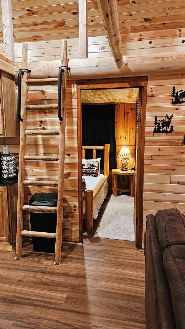 A cozy cabin interior with a wooden ladder, a bed with a patterned pillow, a nightstand with a lamp, and a small table with a decorative item.