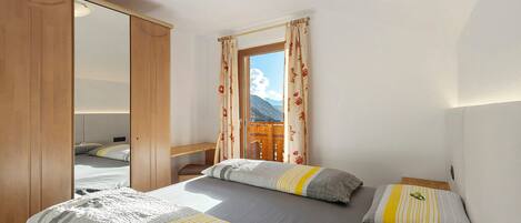 A bedroom with two beds, a wardrobe, a window with curtains, and a view of mountains.