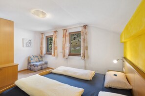 A room with two beds, a sofa, a window with curtains, a radiator, and a wall-mounted light fixture.