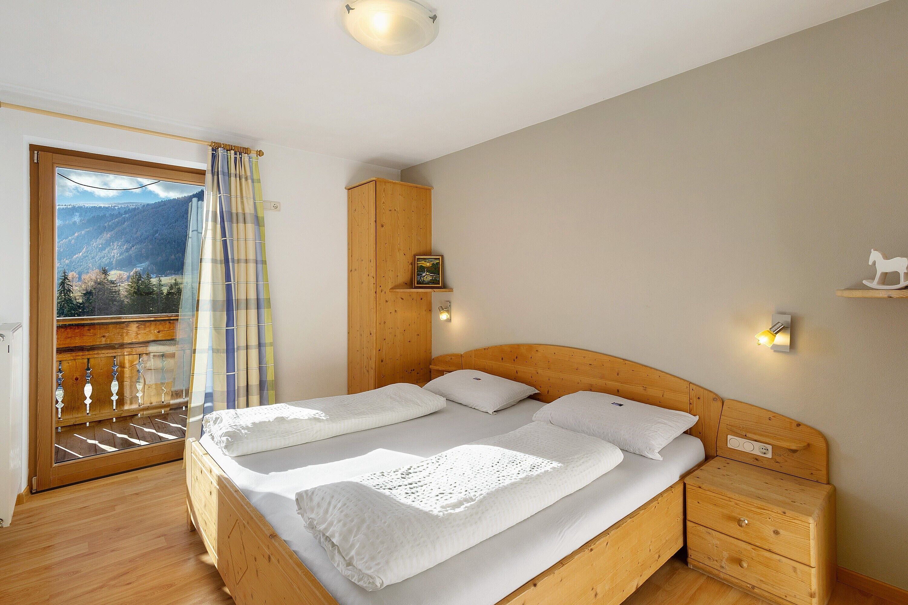 A bedroom with a wooden bed, a nightstand, a wooden wardrobe, a window with a view of mountains, and a balcony with a railing.