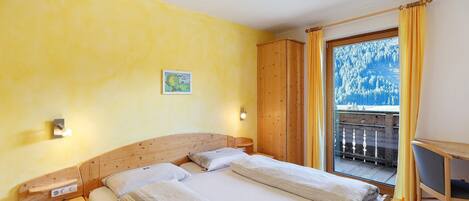 A bedroom with a wooden bed, a desk, and a balcony with a view of trees.