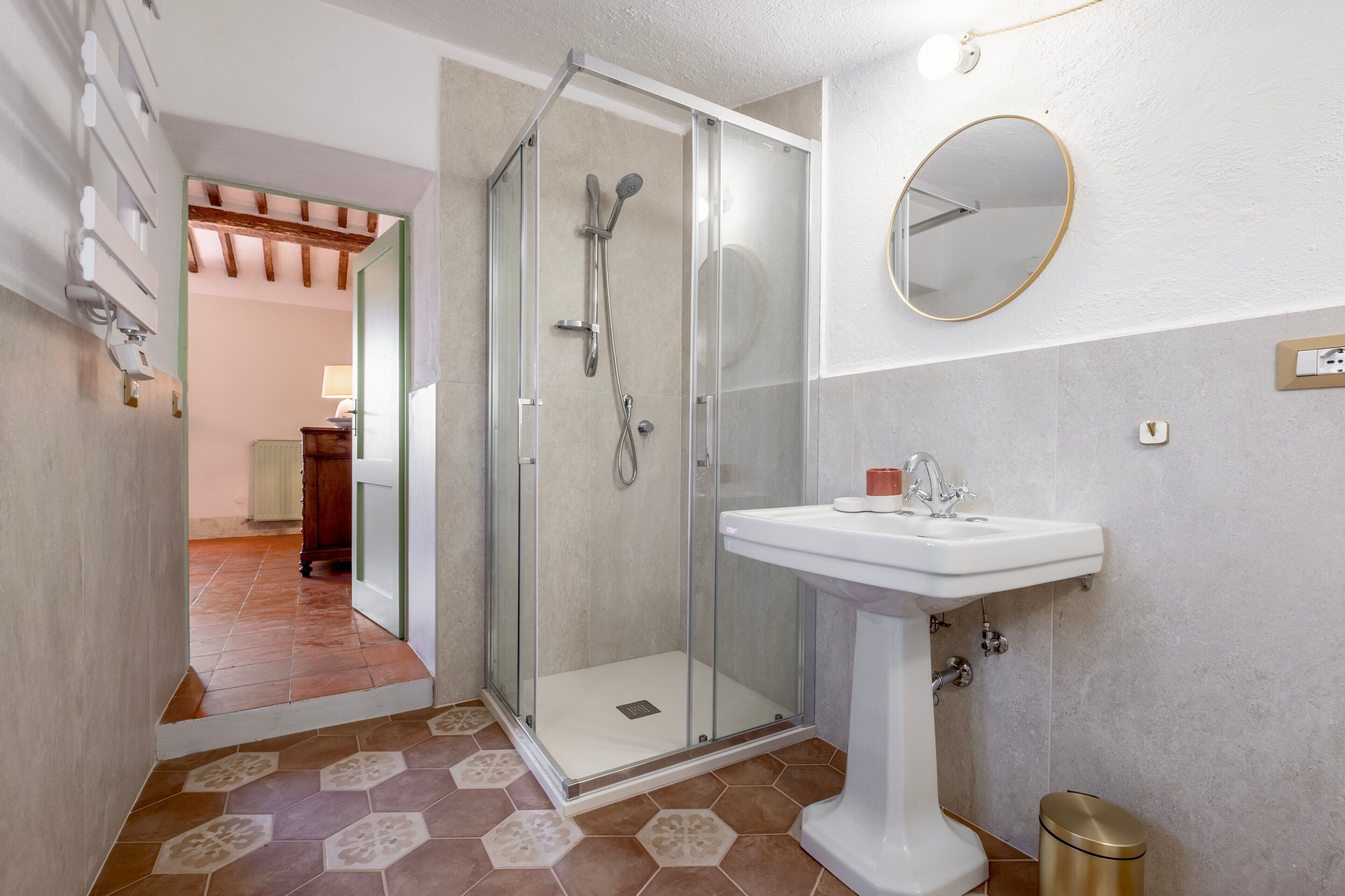 Un baño con ducha envuelta en vidrio, lavamanos pedestal y espejo redondo.