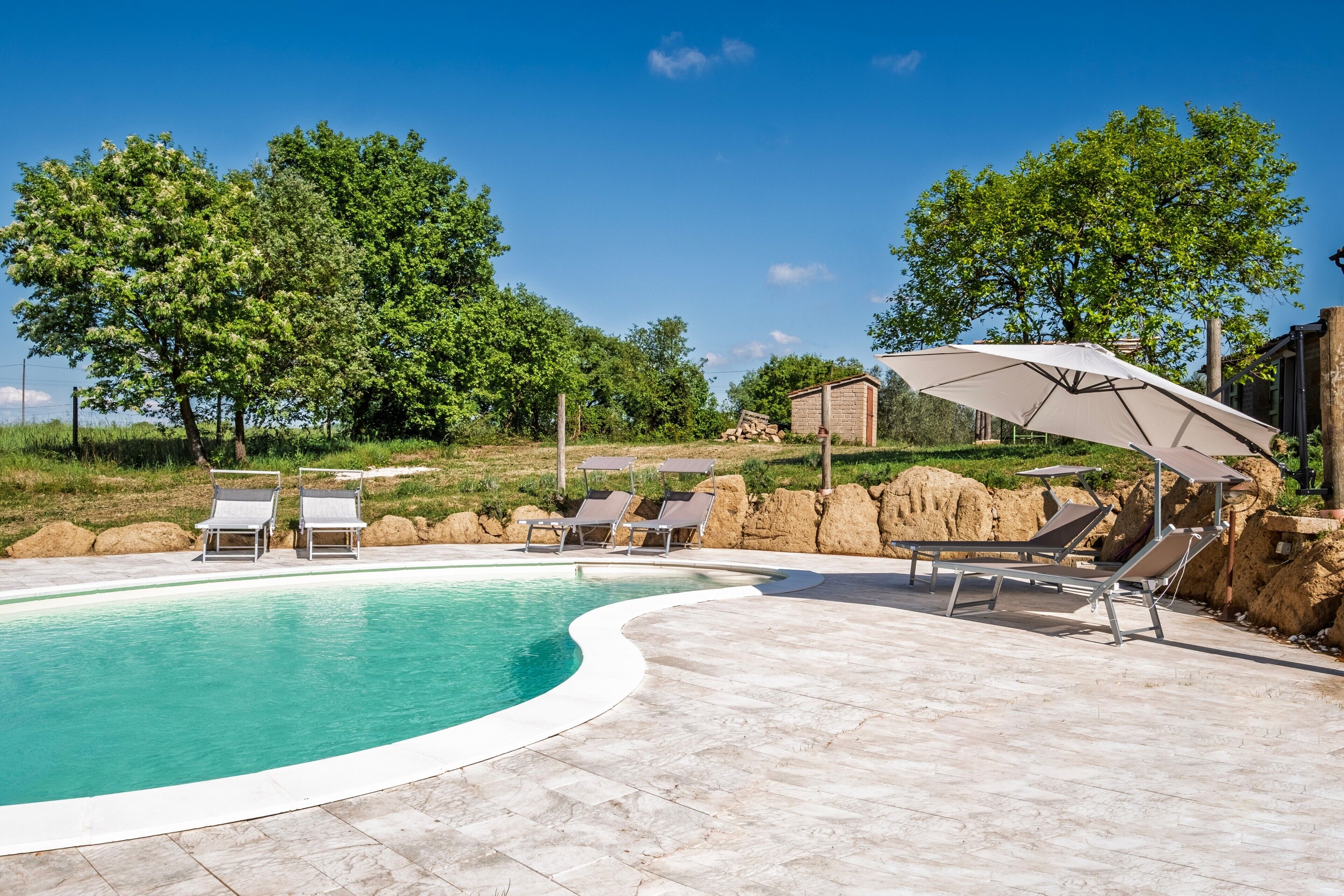 Piscina con sillas de descanso y sombrillas, rodeada por un patio de piedra y árboles.
