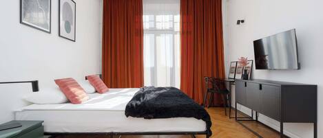 Un dormitorio moderno con cama, escritorio, televisor y ventana con cortinas.