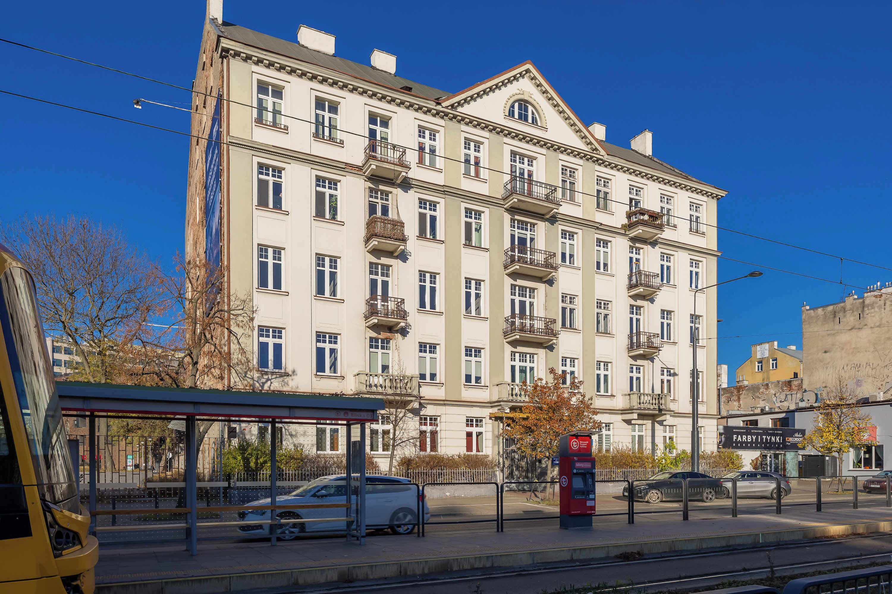 Un edificio a più piani con balconi, una fermata dell'autobus e una linea tramviaria.