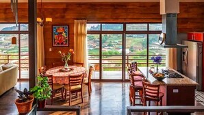 Un comedor amplio con un gran ventanal que ofrece vistas al campo. Hay una mesa redonda con sillas y una pequeña zona de cocina con una barra y taburetes.