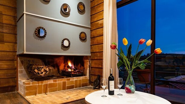 Un interior moderno con chimenea, una mesa redonda con copas de vino y un jarrón de tulipanes, y vistas a través de un gran ventanal.