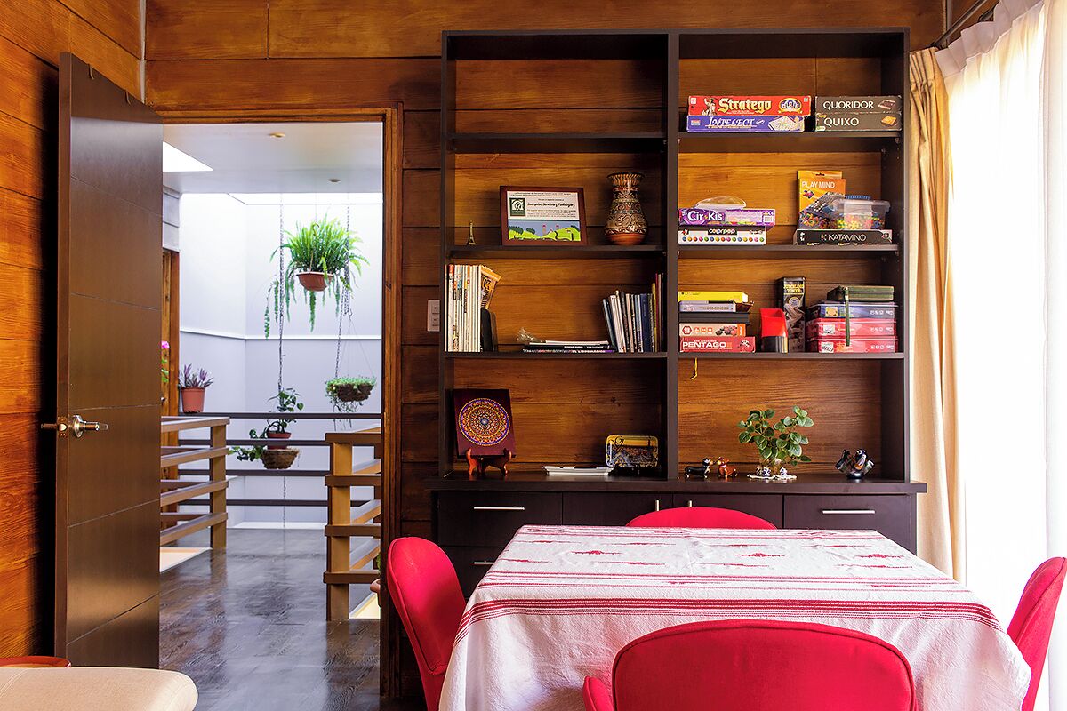Une pièce avec une bibliothèque en bois, une table à manger entourée de chaises rouges et une vue sur un balcon agrémenté de plantes.