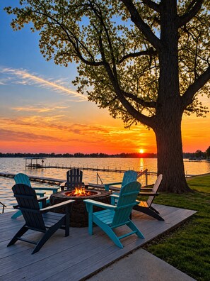 A deck with Adirondack chairs and a fire pit overlooking a body of water at sunset. - Lake Days, Bonfires & Endless Memories (Coldwater)