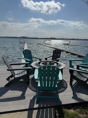 A wooden deck with Adirondack chairs and a circular table, overlooking a calm lake with a dock in the background. - Bella Shores - Stunning Sunsets & Sandy Shoreline (Coldwater)