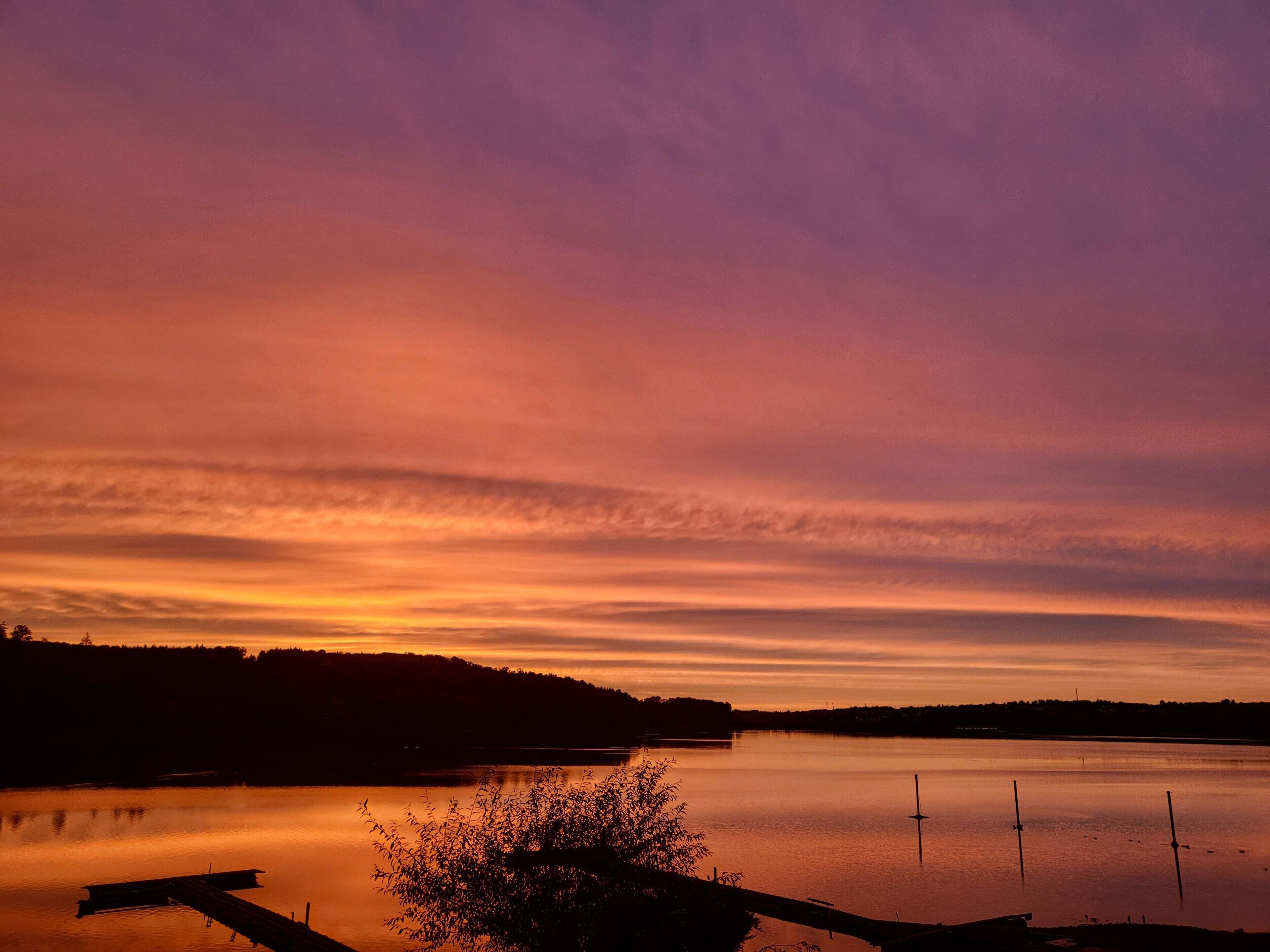 A calm lake at sunset with a dock and trees in the foreground.