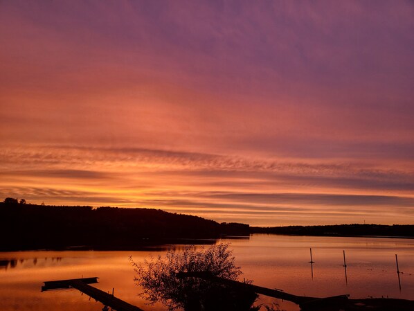 Ein ruhiger See bei Sonnenuntergang mit einem Steg und Bäumen im Vordergrund.