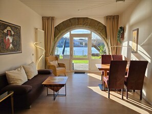 Ein Zimmer mit einer Couch, einem Esstisch mit Stühlen, Blick auf einen See durch ein Rundbogenfenster und einem Gemälde an der Wand.