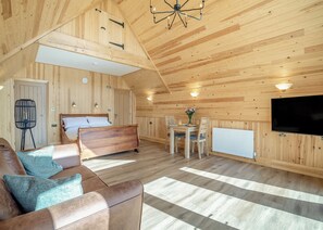 A wooden cabin interior with a bed, sofa, dining table, chairs, a TV, and a radiator.