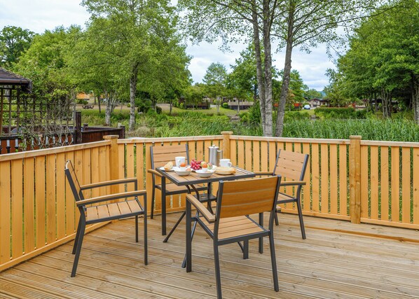 A wooden deck with a table and chairs, surrounded by a wooden fence, overlooking a lush garden with trees and a clear sky.