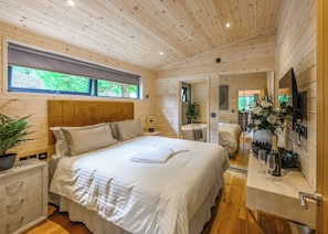 Ein Schlafzimmer einer Holzhütte mit Bett, Nachttisch und Fenster mit Blick auf Grünflächen.