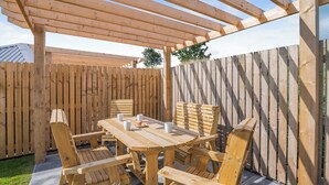 En pergola av trä med ett bord och stolar, ett trästaket och ett träd i bakgrunden.