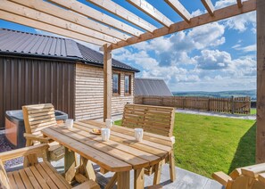 A wooden outdoor seating area with a table and chairs, a stone bench, and a view of a building with a metal roof. - 1 bedroom accommodation in Ripponden (Ripponden)