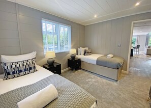 Een slaapkamer met twee bedden, een raam met zonwering, een nachtkastje met een lamp en een deur die leidt naar een andere kamer.